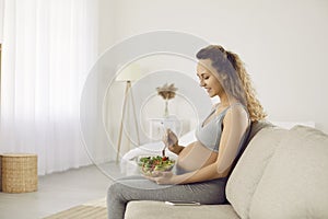 Young pregnant woman sitting on couch and eating bowl of healthy vegetable salad