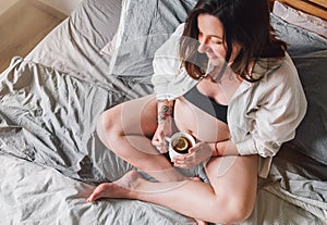 Young pregnant woman sitting with cross-legged on the bed with cup of tea in bedroom e. Woman's health,