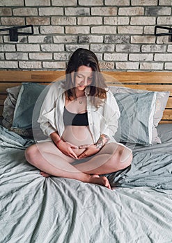 Young pregnant woman sitting with cross-legged tender touching belly on the bed in bedroom in the early morning time.