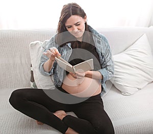 Young pregnant woman sitting on the sofa and reading a book