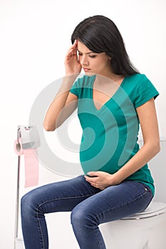 Young pregnant woman sitting in toilet. girl holding head and belly feeling headache