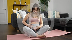 Young pregnant woman sitting on yoga mat touching belly at home