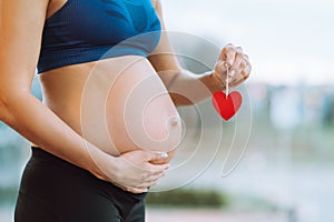 Young pregnant woman in sportswear holding red heart hanging on stick in hand and caressing belly.