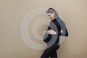 Young pregnant woman standing against beige background looking at her tummy