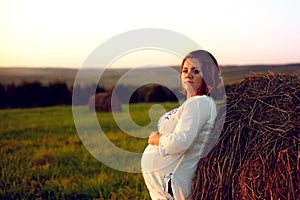 Young Pregnant Woman Standing by the Haystack at Sunset and Embracing her Belly. 8 Month Pregnancy.