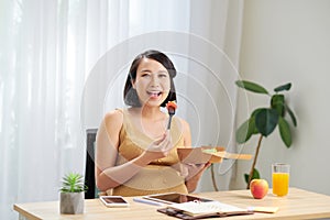 A young pregnant woman standing in home office and using tablet pc. Breakfast time