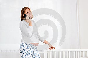 A young pregnant woman is standing in the nursery, leaning on a baby cradle and chatting on her smartphone. Concept of modern