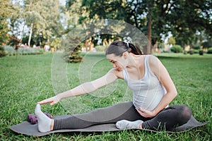 Young pregnant woman stretching outisde in park. She reach feet with one hand on yoga mate. Model keep another hand on
