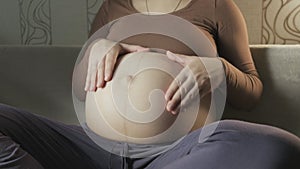 young pregnant woman stroking big belly on couch, making heart shape with hands