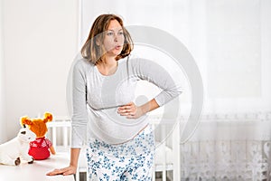 A young pregnant woman suffers from pain and holds her hand on her belly. Children`s room in the background. The concept of healt