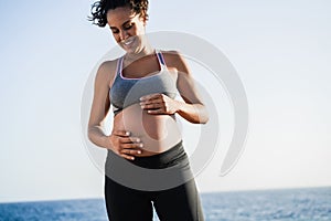 Young pregnant woman touching her belly while doing workout routing outdoor on the beach - Sport exercises and maternity concept