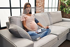 Young pregnant woman using blood pressure monitor sitting on the sofa afraid and shocked with surprise and amazed expression, fear