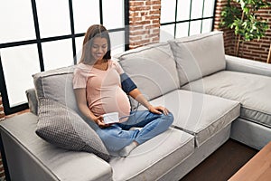Young pregnant woman using blood pressure monitor sitting on the sofa smiling with a happy and cool smile on face