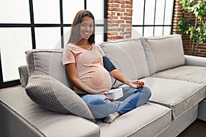 Young pregnant woman using blood pressure monitor sitting on the sofa smiling and laughing hard out loud because funny crazy joke