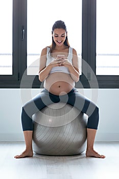 Young pregnant woman using her mobile phone while doing relaxation exercise with fitness ball at home