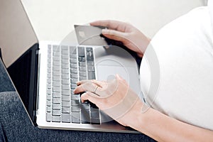 Young pregnant woman using laptop for online shopping and holding credit card to making payment sit on sofa at home.