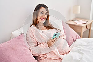Young pregnant woman using smartphone sitting on bed at bedroom