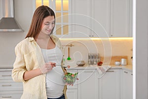 Young pregnant woman with vegetable salad in kitchen, space for text. Healthy eating