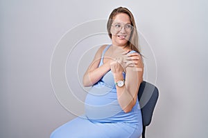 Young pregnant woman wearing band aid for vaccine injection pointing thumb up to the side smiling happy with open mouth