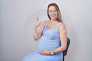 Young pregnant woman wearing band aid for vaccine injection smiling happy and positive, thumb up doing excellent and approval sign
