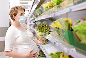 Young pregnant woman wearing disposable medical face mask choosing fresh salad in supermarket during coronavirus outbreak