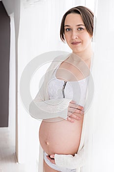 Young pregnant woman in white negligee standing at the window, selective focus