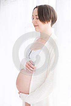 Young pregnant woman in white negligee standing at the window, selective focus