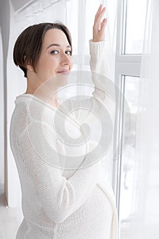 Young pregnant woman in white negligee standing at the window, selective focus