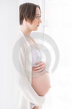 Young pregnant woman in white negligee standing at the window, selective focus