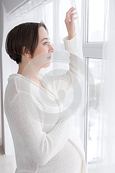 Young pregnant woman in white negligee standing at the window, selective focus