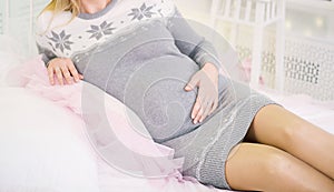 Young pregnant woman in winter dress lying on bed at home and touching her belly, close up. Pregnancy and expectation concept