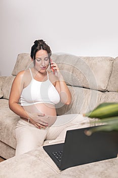 A young pregnant woman works with a laptop on her couch at home, while talking on the phone and touching her belly.