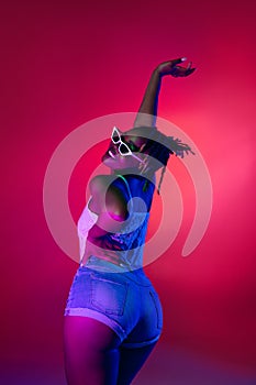 Young pretty African girl in sunglasses dancing on heels isolated on gradient studio background in neon light.