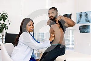 Young pretty African woman doctor examining African man patient with stethoscope in medical office