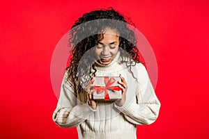 Young pretty african woman smiling and holding gift box on red studio background