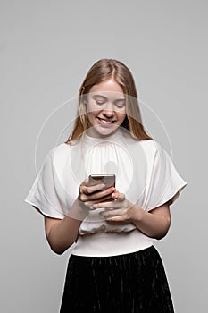 Young pretty blond girl with long hair smiling and using cell phone isolated on gray background. Modern communication