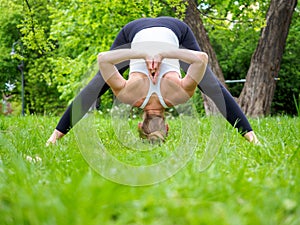 young pretty blond woman practicing yoga outdoor in the park in a sunny summer day, standing in prasarita padottanasana pose