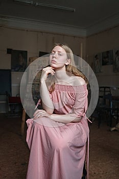 Young pretty blonde girl in pink dress propping her head with arm in art studio, looking questioningly into camera