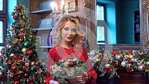 A young pretty blonde girl in a red dress decorates the Christmas tree with Christmas toys