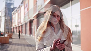 Young pretty blonde lady walking down the lonesome street in the city center, using her smartphone for communication.