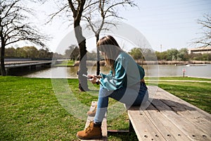 A young, pretty, blonde woman is sitting at a wooden table in the park, looking at her mobile phone and smoking a cigarette. In