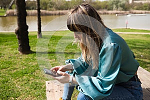 A young, pretty, blonde woman is sitting at a wooden table in the park, looking at her mobile phone and smoking a cigarette. In