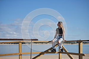 Young, pretty, blonde woman, wearing a black top, ripped jeans and tattoos, sitting on a railing, looking at infinity with the sea