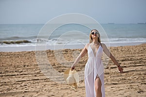 Young, pretty, blonde woman in a white dress, sunglasses and hat, with open arms, grateful, relaxed and happy, on the beach.