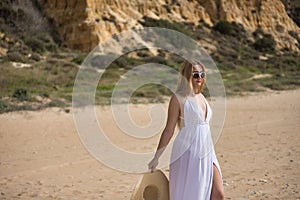 Young, pretty, blonde woman in a white dress, sunglasses and hat, walking on the beach. Concept beach, summer, vacation