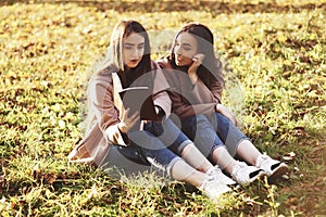 Young pretty brunette twin girls sitting on the grass with legs slightly bent in knees and looking in a brown book