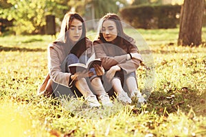 Young pretty brunette twin girls sitting on the grass with legs slightly bent in knees and reading in a brown book