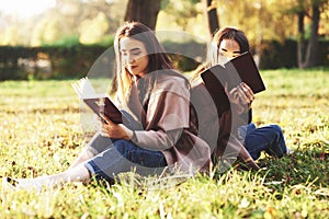 Young pretty brunette twin sisters sitting back to back on the grass with legs slightly bent in knees with brown books