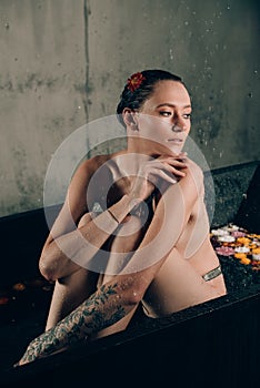 Young pretty brunette woman in bath with water and flowers. Naked model during an unusual photo shoot in a black bath