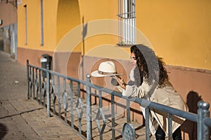 Young, pretty, brunette woman with curly hair and beige coat and hat, leaning on a railing looking at her hat thoughtful and